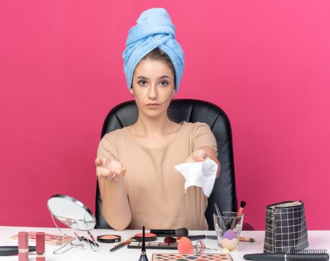 young-beautiful-girl-sits-table-with-makeup-tools-wrapped-hair-towel-holding-out-napkin-isolated-pink-wall